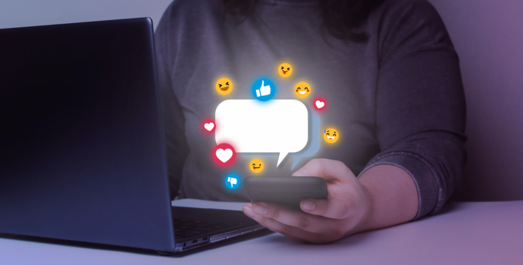 Persona usando celular junto a laptop con burbuja de diálogo y emojis flotando